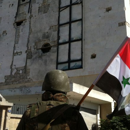 A Syrian army's soldier a national flag featuring Syria's President Bachar al-Assad in front of a building left in ruins with on June 5, 2013 in the city of Qusayr in Syria's central Homs province, after the Syrian government forces seized total control of the city and the surrounding region. The Syrian army ousted rebels from the strategic town of Qusayr after a blistering 17-day assault led by Hezbollah fighters, scoring a major battlefield success in a war that has killed at least 94,000 people.   AFP PHOTO / STR        (Photo credit should read -/AFP/Getty Images)